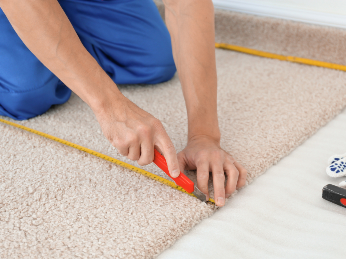 A Room Measurement Guide: Basics for Better Flooring Estimates 2 Person cutting beige carpet with a utility knife along a yellow measuring tape, using it as a measurement guide. Tools and carpet padding are visible nearby.