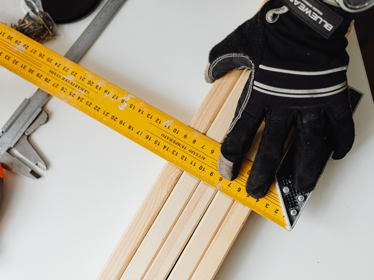 A Room Measurement Guide: Basics for Better Flooring Estimates 4 A gloved hand holds a yellow measuring tape and square as a measurement guide against several wooden planks on a white surface, with tools and screws nearby.
