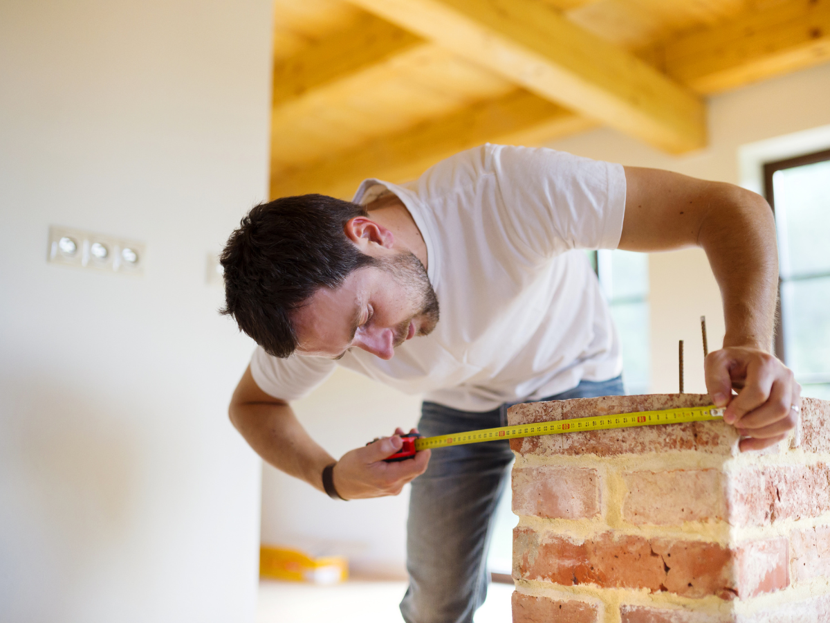 A Room Measurement Guide: Basics for Better Flooring Estimates 5 Man in a white t-shirt uses a tape measure as a measurement guide on top of a brick structure inside a house with wooden beams.