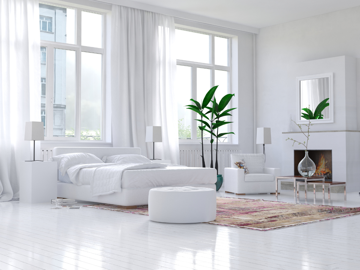 Cozy & Stylish Bedroom Flooring Ideas 5 Bright, modern bedroom with white walls, large windows, a bed, armchair, fireplace, green plants, and a rug that enhances the sleek white bedroom flooring.