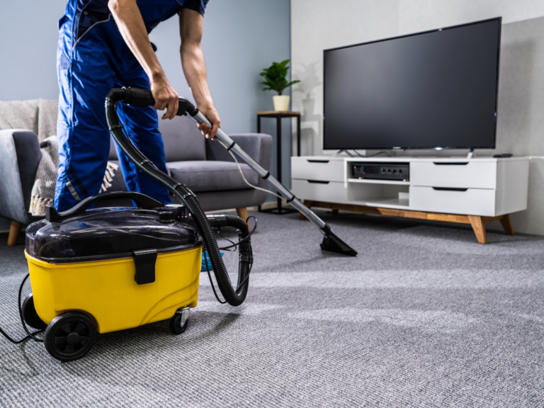 A person is performing carpet cleaning on a gray carpet in a living room with a yellow industrial vacuum cleaner; a TV and console can be seen in the background.