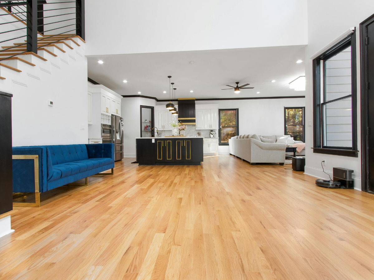 Frequently Asked Questions About Flooring: Answers to Your Top Questions 2 Modern open-concept living area with hardwood floors, blue sofa, black kitchen island, stainless appliances, and neutral sectional sofa. Large windows and staircase are visible—please refer to our Frequently Asked Questions for more details on design options and amenities.