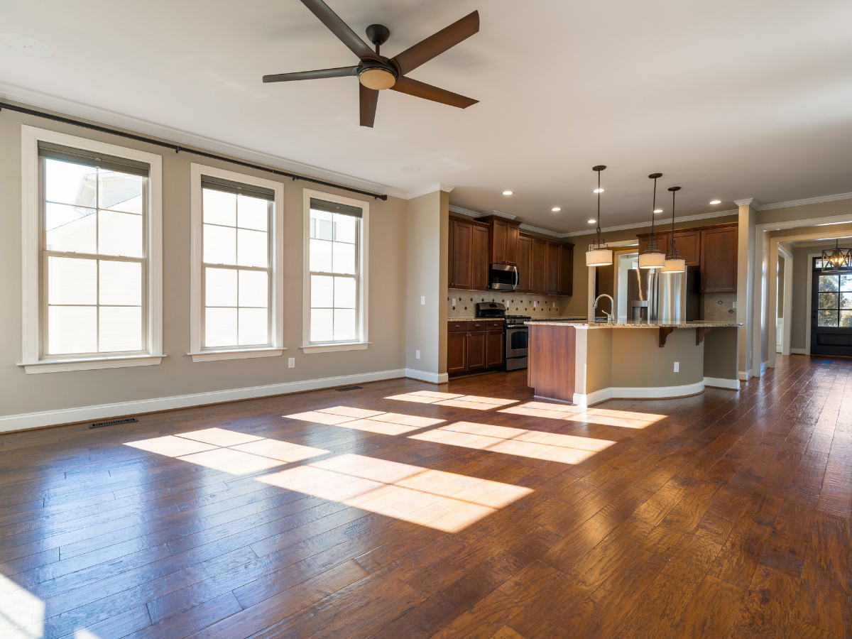 Frequently Asked Questions About Flooring: Answers to Your Top Questions 5 Sunlit empty living room with hardwood floors, ceiling fan, and an open kitchen featuring brown cabinets, stainless steel appliances, and pendant lighting—perfect for relaxing after a long day or answering Frequently Asked Questions about your ideal home.