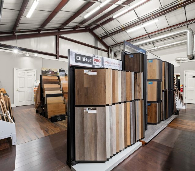 A showroom displays various samples of wood and laminate flooring arranged on racks under a high, industrial-style ceiling, offering inspiration for your next laminate flooring installation.